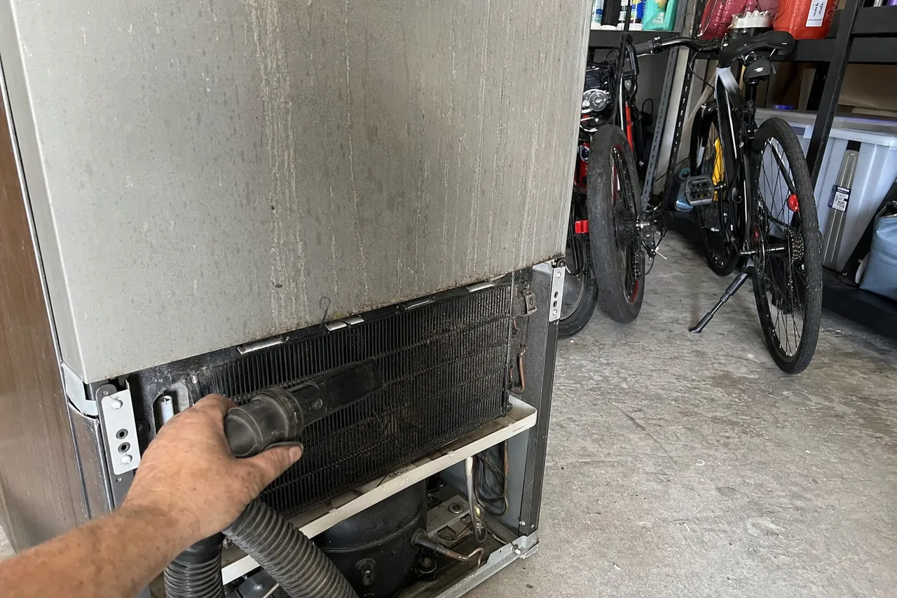 A refrigerator pulled forward in a Florida garage with its lower grille off and the condenser exposed, bicycles and shelving stacked behind it.