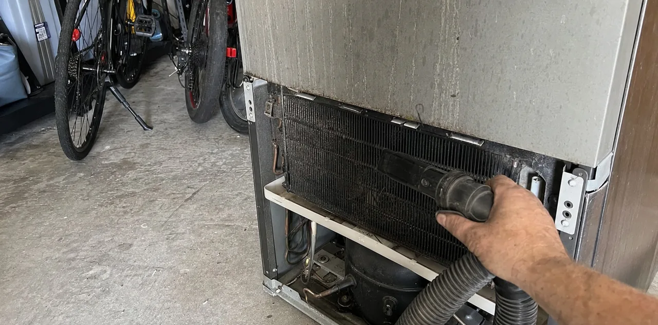 A vacuum hose worked along the base of a condenser coil, the compressor visible in the shadow behind it.