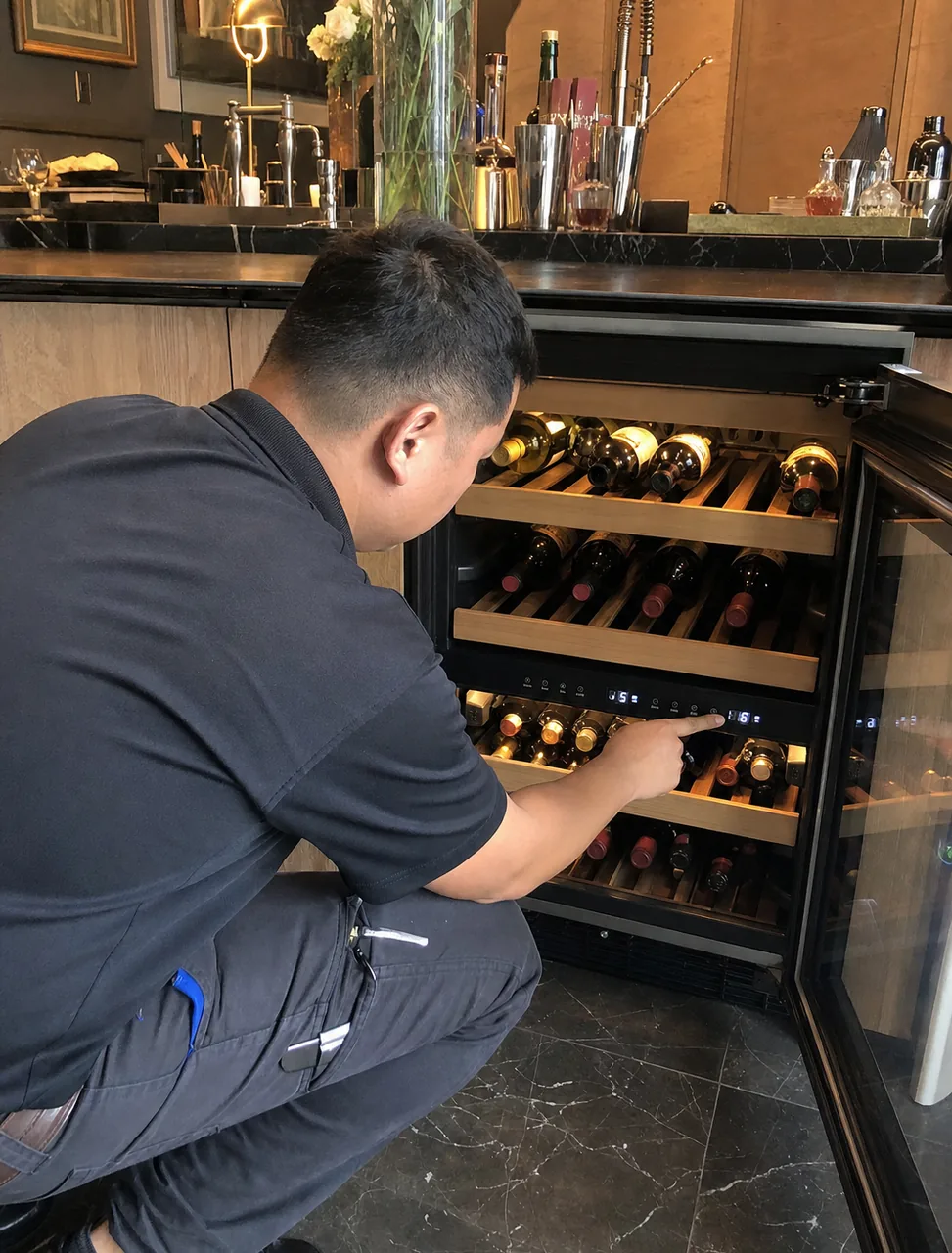 A technician checking a two-zone wine cabinet, bottles racked on wooden runners above and below the divider.